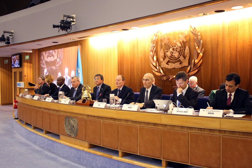 Réunion de responsables assis autour d’une table dans une salle de conférence ornée de l’emblème des Nations unies et d’un drapeau bleu.