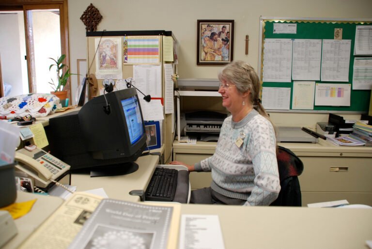 Une personne travaille à un bureau devant un ordinateur, entourée de documents, d’un téléphone, d’imprimantes et de tableaux d’affichage.
