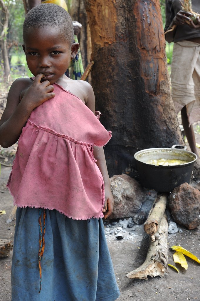 Une enfant se tient devant une marmite posée sur un feu de bois, dans un environnement extérieur arboré.