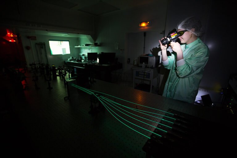 Personne en tenue de laboratoire utilisant un appareil optique dans une salle sombre, avec des faisceaux laser verts sur une table expérimentale et du matériel scientifique en arrière-plan.