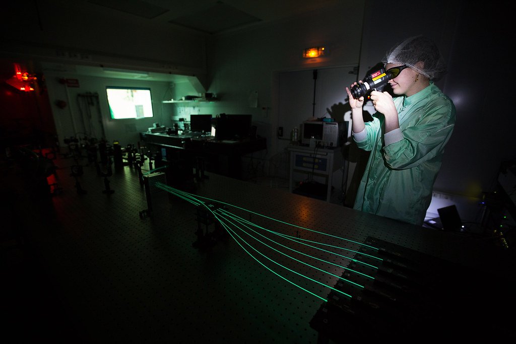 Personne en tenue de laboratoire utilisant un appareil optique dans une salle sombre, avec des faisceaux laser verts sur une table expérimentale et du matériel scientifique en arrière-plan.