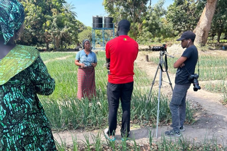 Une équipe filme une femme dans un champ cultivé, avec une caméra installée sur trépied et des réservoirs d’eau en arrière-plan.