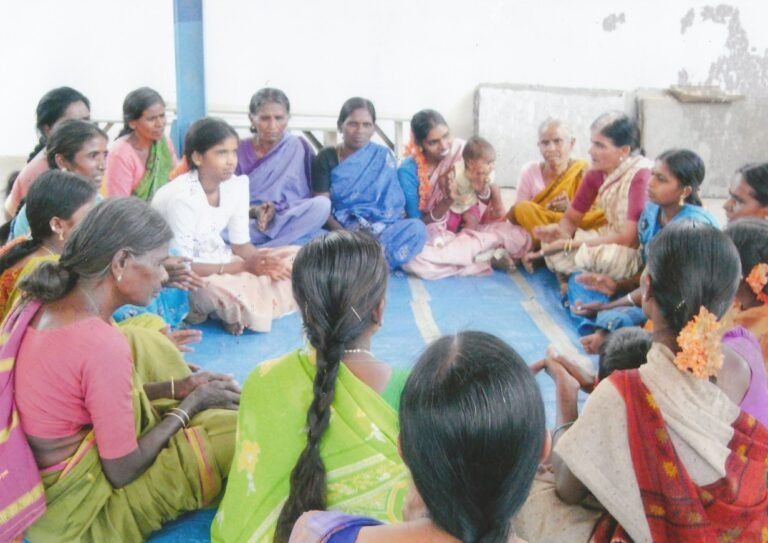 Un groupe de femmes assises en cercle dans une salle, certaines échangeant entre elles, tandis qu’une femme tient un bébé.
