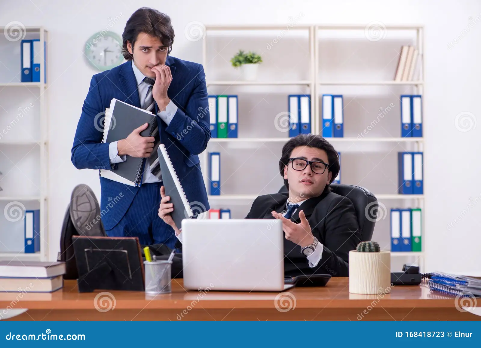 Deux hommes en costume travaillent dans un bureau. L’un est assis derrière un ordinateur portable et l’autre se tient à côté de lui avec des dossiers. Des classeurs sont rangés sur des étagères derrière eux.