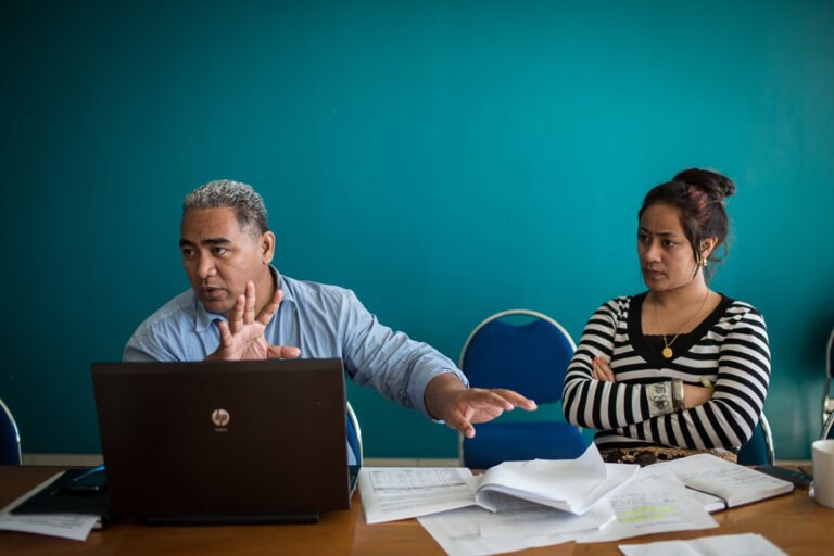 Deux personnes sont assises à une table dans une salle de réunion. Un homme parle en faisant un geste devant un ordinateur portable, tandis qu’une femme l’écoute. Des documents et des cahiers sont posés sur la table.