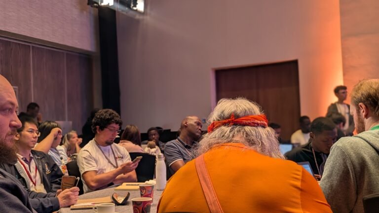 Des personnes réunies autour de tables dans une salle, certaines consultant un ordinateur, un téléphone ou des documents.