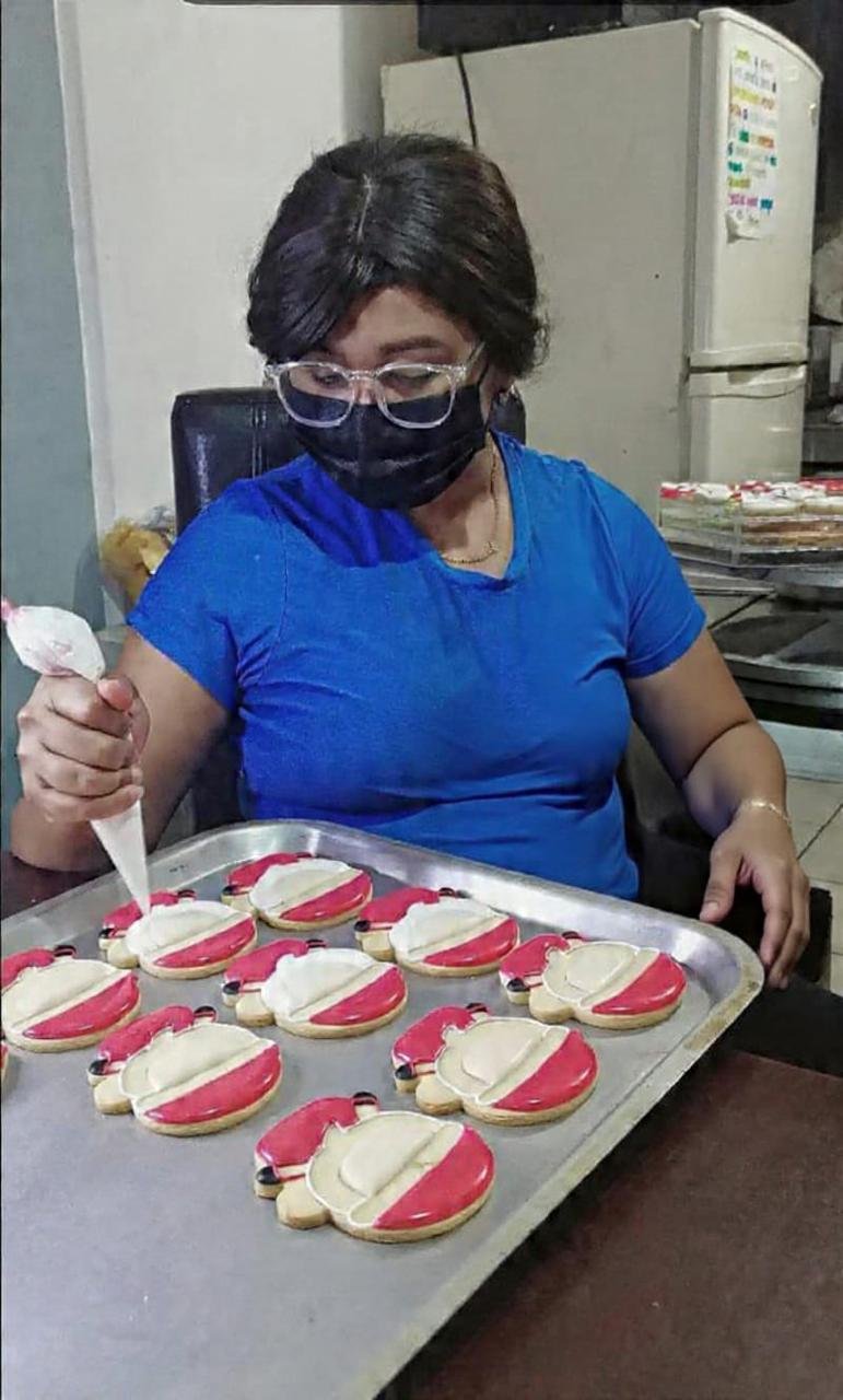Une personne portant des lunettes et un masque décore des biscuits sur une plaque dans un espace de préparation alimentaire.