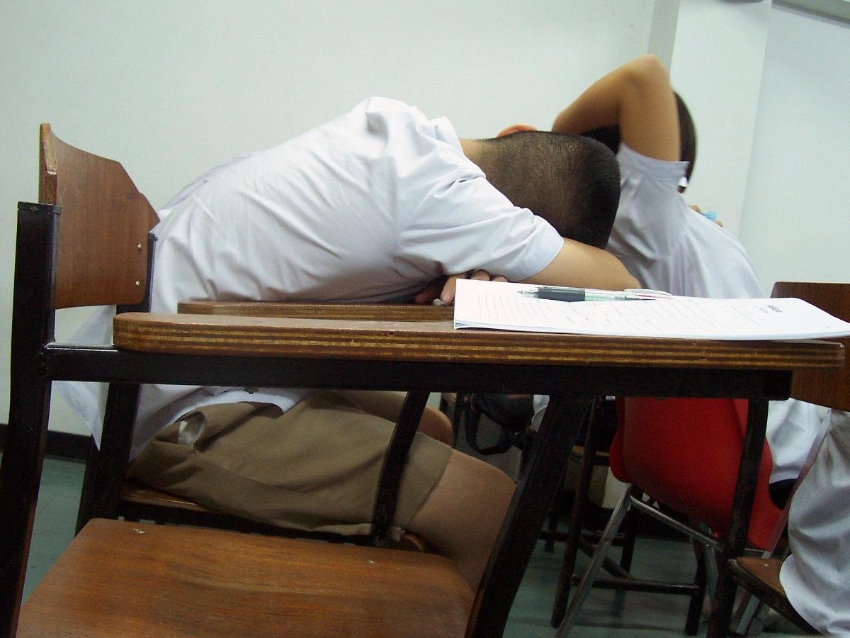 Deux étudiants endormis, la tête posée sur un bureau dans une salle de cours, avec des cahiers et des stylos devant eux.