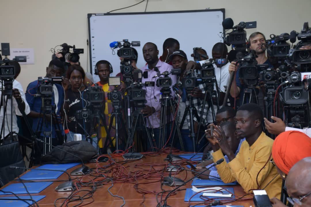 Des journalistes et des cadreurs équipés de caméras et de microphones sont réunis dans une salle autour d’une table couverte de câbles et de matériel audiovisuel.