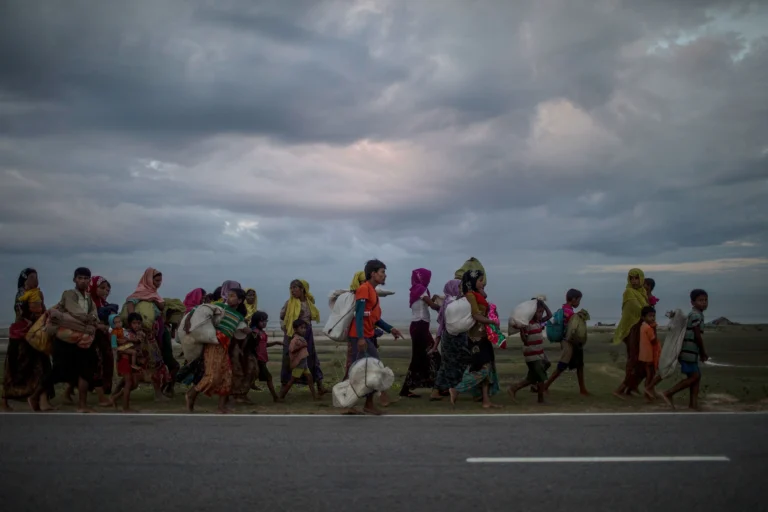 Un groupe de personnes, dont plusieurs enfants, marche avec des sacs et des effets personnels le long d’une route sous un ciel nuageux.