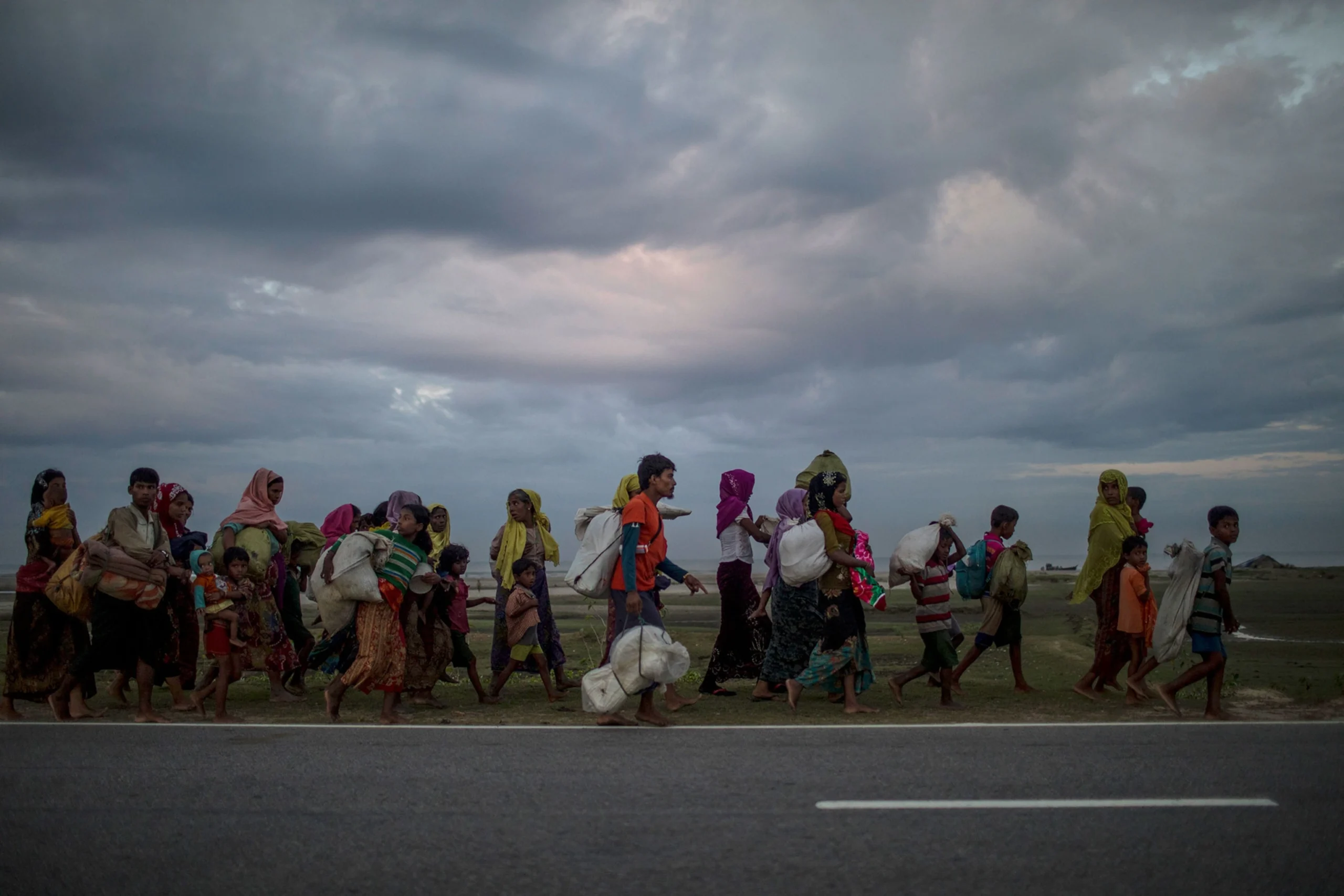 Un groupe de personnes, dont plusieurs enfants, marche avec des sacs et des effets personnels le long d’une route sous un ciel nuageux.