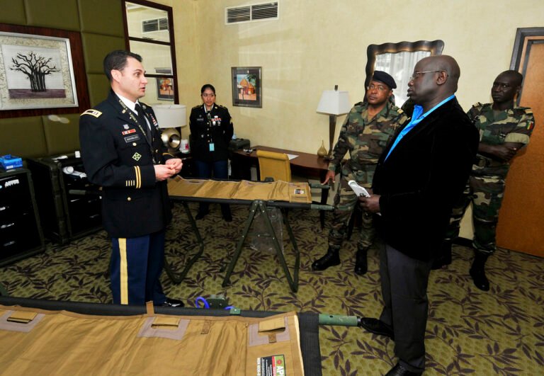 Plusieurs personnes, dont des militaires en uniforme et un homme en costume, échangent dans une salle autour de tables portant du matériel médical ou logistique.