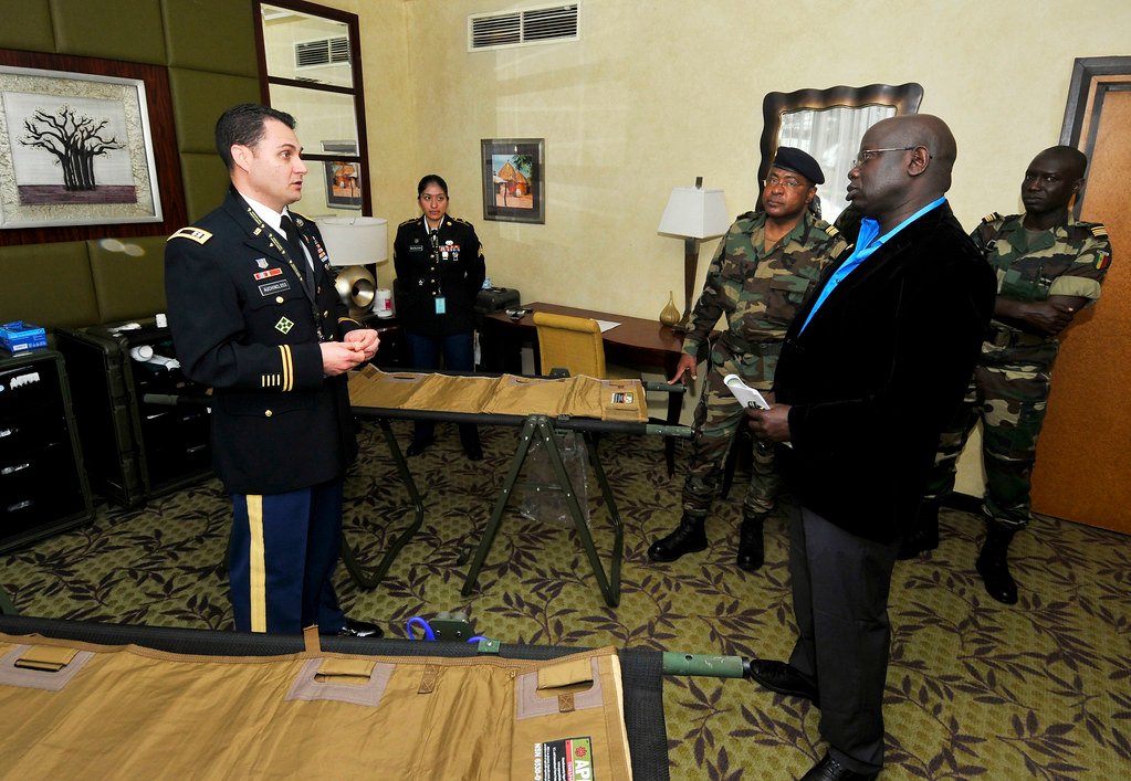 Plusieurs personnes, dont des militaires en uniforme et un homme en costume, échangent dans une salle autour de tables portant du matériel médical ou logistique.