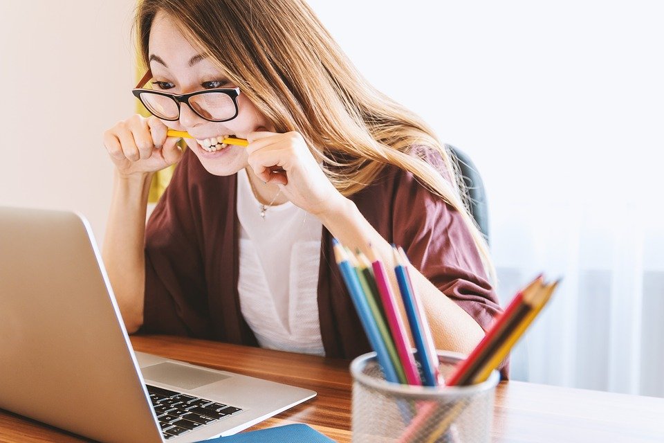 Jeune personne portant des lunettes, assise devant un ordinateur portable et mordant un crayon à un bureau avec un pot de crayons de couleur.