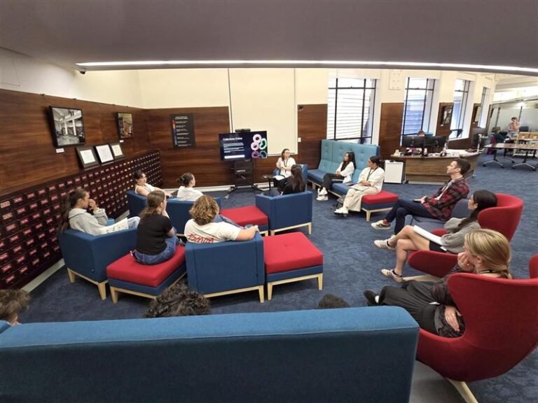 Groupe d’étudiants assis dans un espace intérieur de bibliothèque ou d’établissement universitaire, près d’étagères et d’un écran d’information.