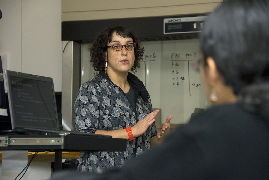 Une femme présente un sujet devant un tableau dans une salle équipée d’un ordinateur et de matériel de laboratoire, tandis qu’une autre personne est visible au premier plan.