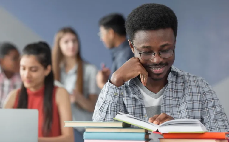 Un jeune homme assis à une table consulte un livre ouvert posé sur une pile de livres. À l’arrière-plan, d’autres jeunes sont assis ou discutent, dont une personne devant un ordinateur portable.