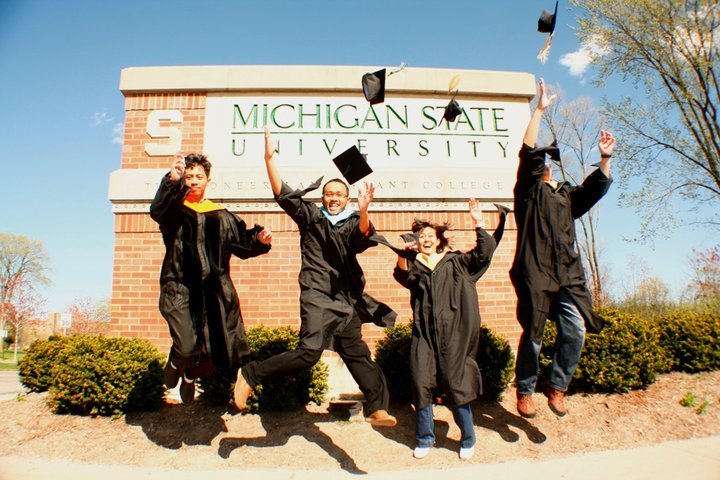 Quatre diplômés en toge lancent leurs chapeaux devant un panneau de la Michigan State University.