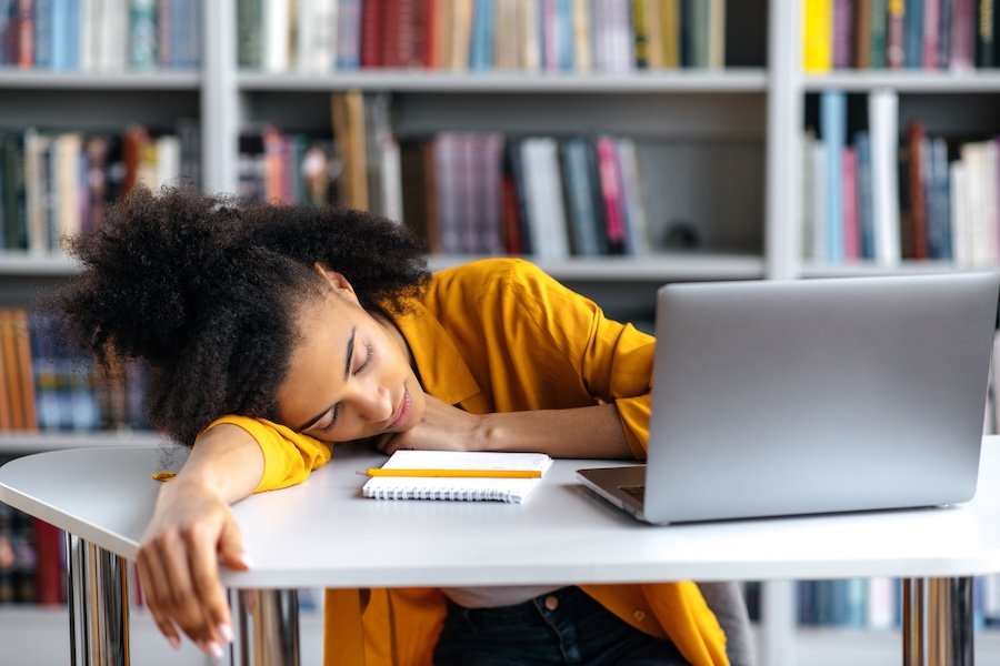 Une jeune personne vêtue d’un vêtement jaune dort, la tête posée sur son bras, devant un ordinateur portable et des cahiers, dans une bibliothèque.