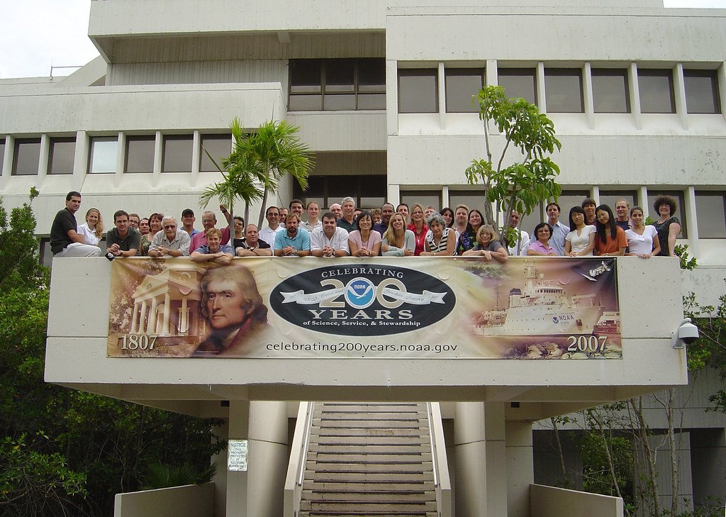 Groupe de personnes rassemblées sur une passerelle devant un bâtiment, avec une grande bannière NOAA accrochée à la balustrade.