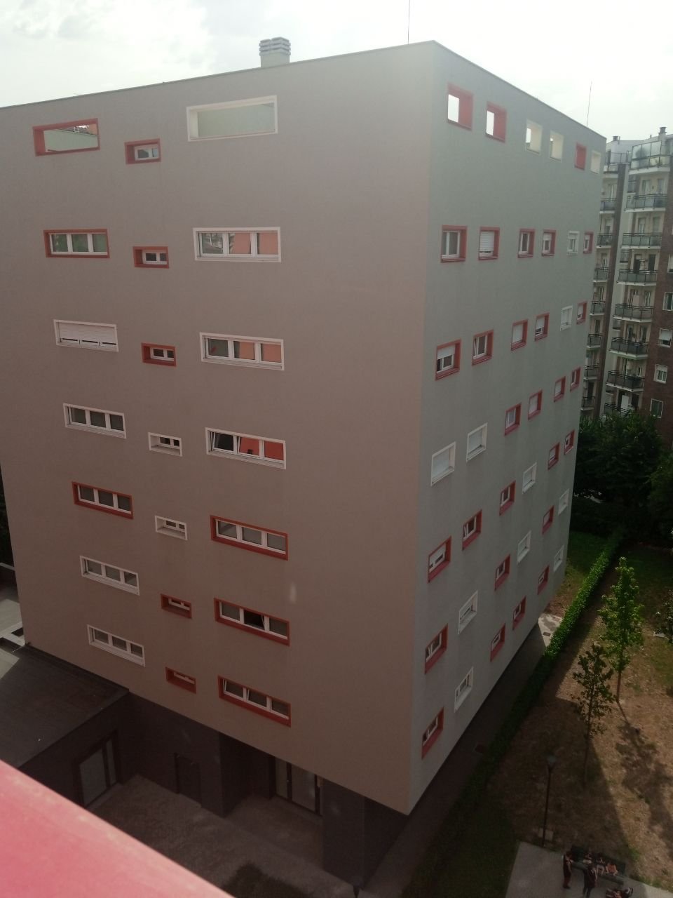Vue extérieure d’un bâtiment résidentiel à plusieurs étages, aux façades grises et roses, entouré d’autres immeubles et d’un espace arboré.