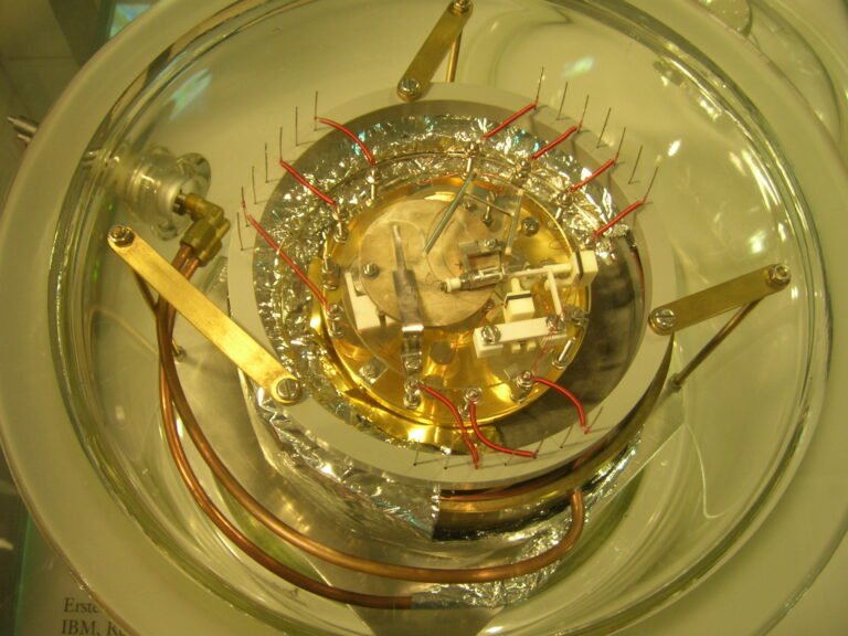 Dispositif expérimental scientifique sous une cloche transparente, entouré de câbles, de pièces métalliques et de composants techniques.