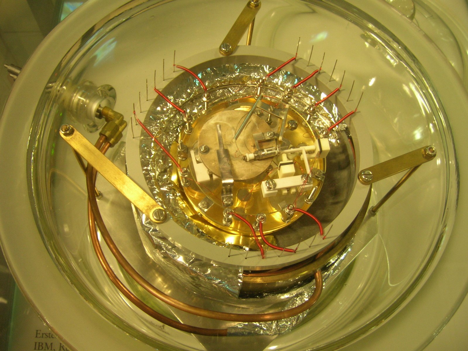 Dispositif expérimental scientifique sous une cloche transparente, entouré de câbles, de pièces métalliques et de composants techniques.