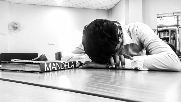 Une personne dort la tête posée sur une table dans une bibliothèque, à côté de plusieurs livres, dont un ouvrage portant l'inscription « MANDELA ».