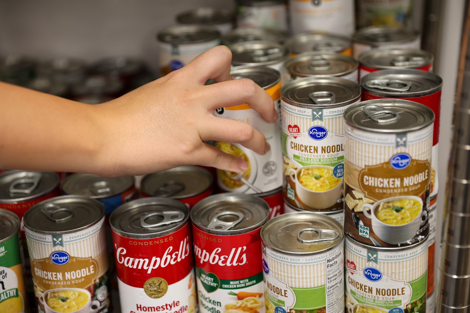 Une main prend une boîte de soupe parmi de nombreuses boîtes de conserve empilées sur des étagères.