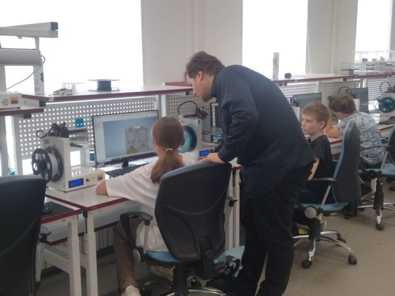 Dans un atelier équipé d’ordinateurs et de matériel technique, un adulte se penche auprès d’une élève assise devant un écran ; deux autres élèves travaillent à proximité.