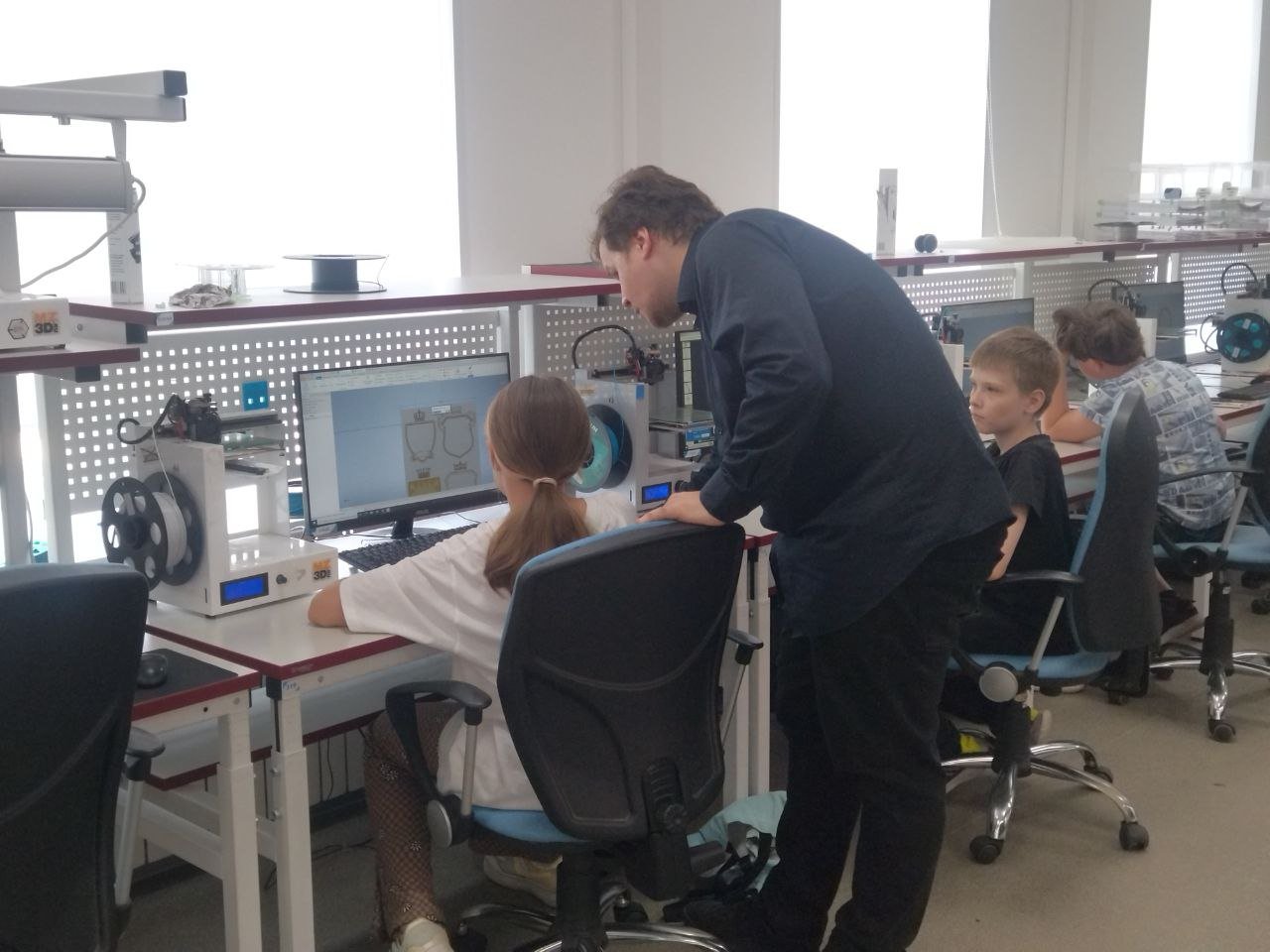 Dans un atelier équipé d’ordinateurs et de matériel technique, un adulte se penche auprès d’une élève assise devant un écran ; deux autres élèves travaillent à proximité.