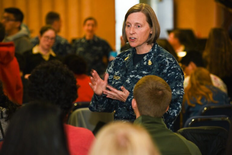 Une femme en uniforme parle en faisant des gestes devant un groupe de personnes assises.