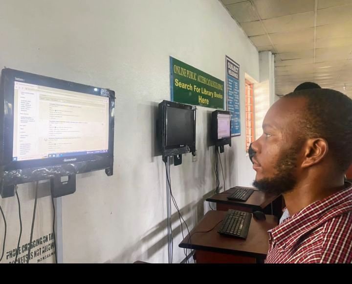 Un étudiant assis devant un ordinateur dans une bibliothèque, avec des postes informatiques et une affiche indiquant la recherche de livres au catalogue.