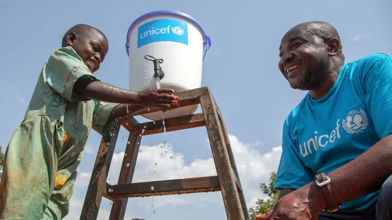 Un enfant se lave les mains sous un robinet fixé à un réservoir surélevé, tandis qu’un homme portant un tee-shirt UNICEF le regarde.