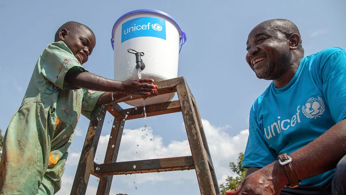Un enfant se lave les mains sous un robinet fixé à un réservoir surélevé, tandis qu’un homme portant un tee-shirt UNICEF le regarde.