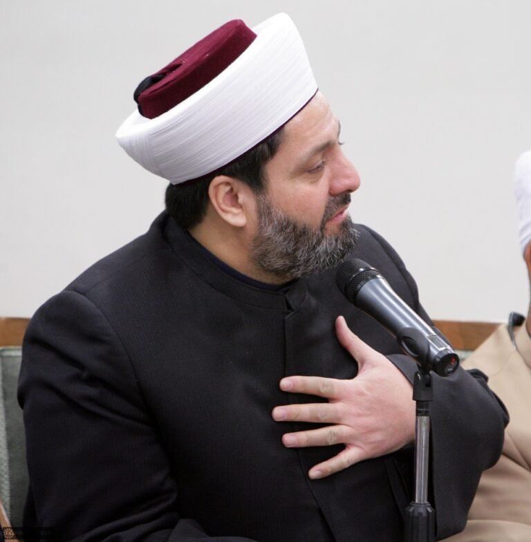 Un homme en tenue sombre et coiffé d’un turban parle dans un microphone, la main posée sur la poitrine ; une autre personne est partiellement visible à côté de lui.