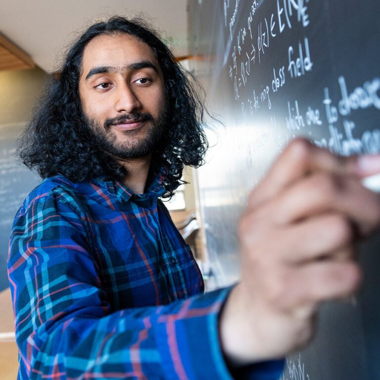 Personne aux cheveux longs écrivant des équations sur un tableau noir dans une salle de cours ou de recherche.