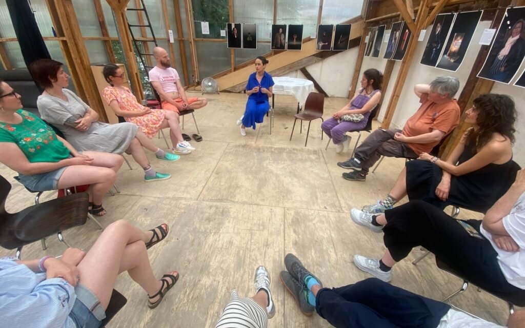 Un groupe d’adultes assis en cercle dans une salle ; une femme en bleu parle au centre tandis que les autres l’écoutent.