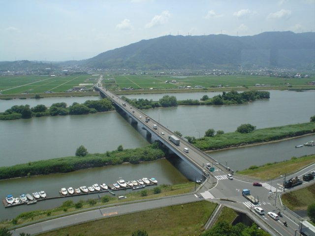 Vue aérienne d’un pont routier traversant un large cours d’eau vers une zone de champs. Des véhicules circulent sur le pont et aux intersections voisines.