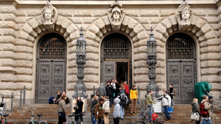 Des personnes se tiennent sur les marches devant l’entrée ornée d’un grand bâtiment universitaire ; des vélos sont stationnés au premier plan.