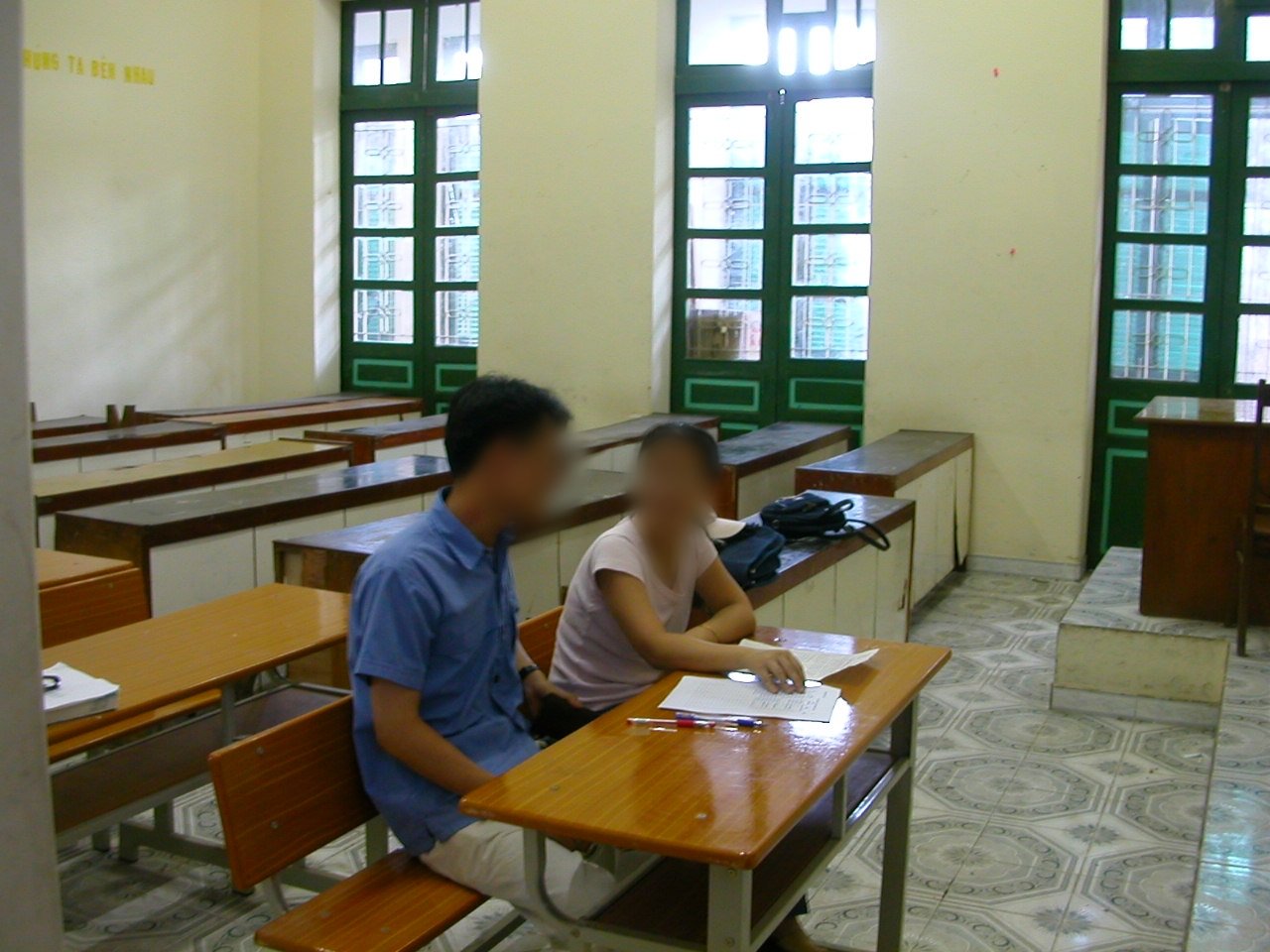Deux personnes sont assises à une table dans une salle de classe, avec des feuilles et un stylo devant elles.