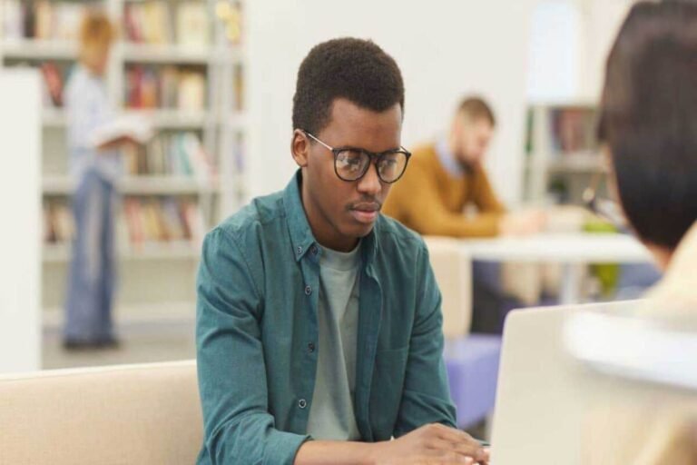 Un étudiant portant des lunettes travaille sur un ordinateur dans un espace d’étude, tandis que d’autres personnes sont assises à des tables et qu’une bibliothèque se trouve en arrière-plan.