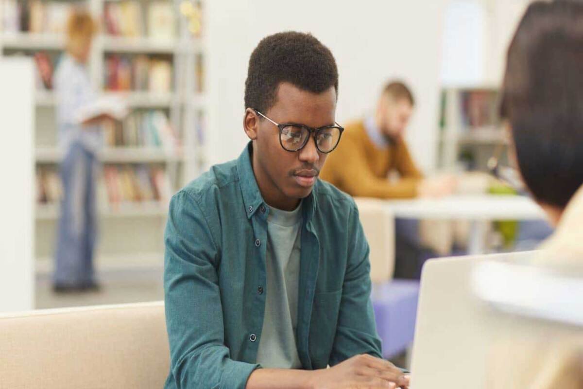 Un étudiant portant des lunettes travaille sur un ordinateur dans un espace d’étude, tandis que d’autres personnes sont assises à des tables et qu’une bibliothèque se trouve en arrière-plan.