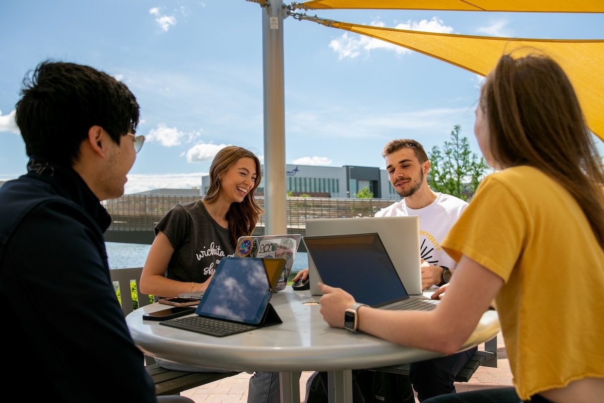 Quatre étudiants travaillent ensemble sur des ordinateurs et des tablettes autour d’une table extérieure, sur un campus universitaire.