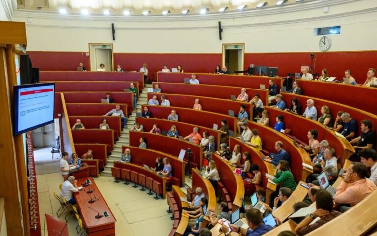 Des personnes sont assises dans un grand amphithéâtre universitaire aux rangées courbes, certaines devant des ordinateurs portables.